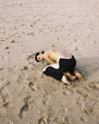 Person kneeling on a sandy beach covered in footprints, wearing dark trousers with white side panels