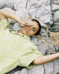 A model lying on rocky terrain holding a stone near her eye, wearing a green embroidered shirt and blue hoop earrings.