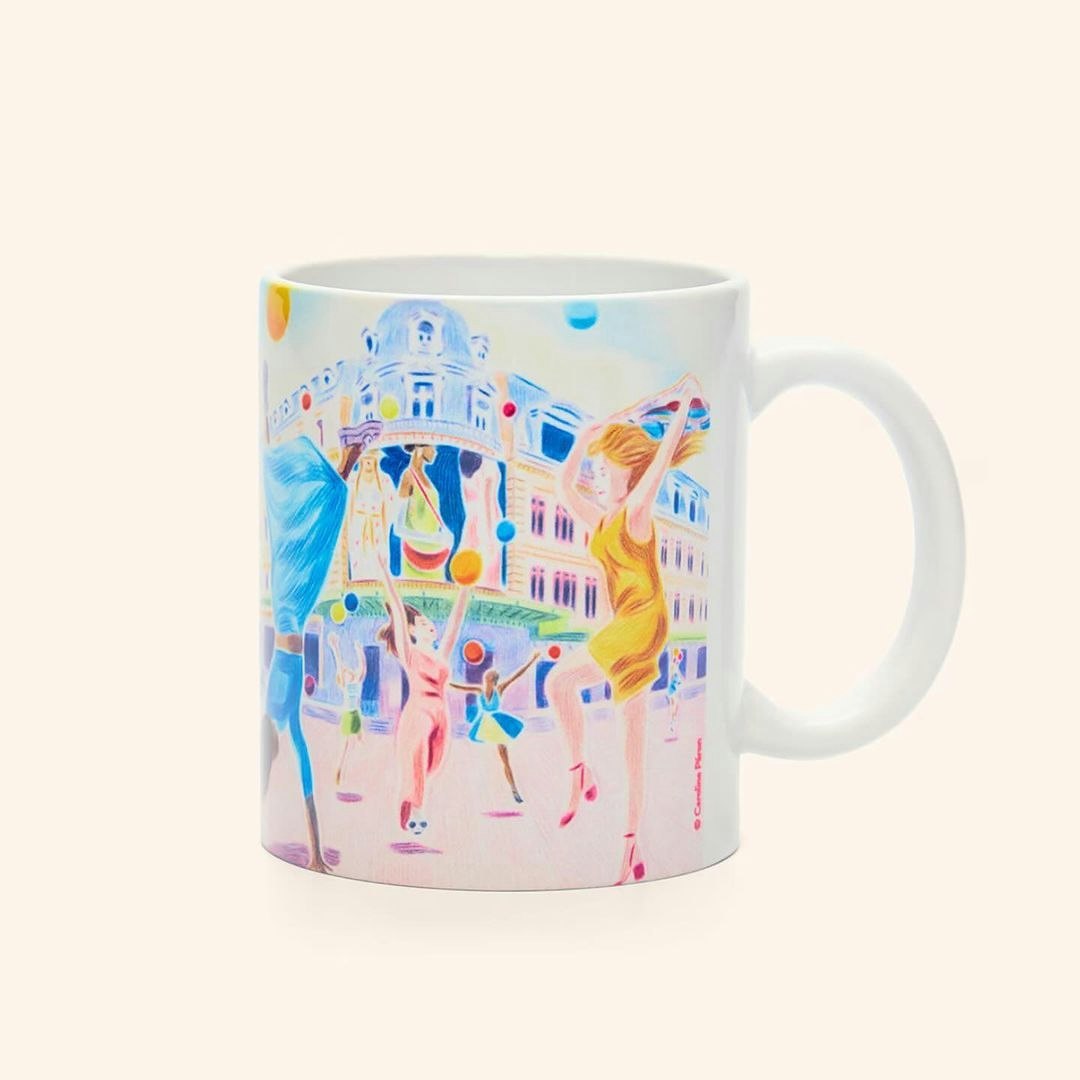 Artistic rendering on a mug: illustration of figures in yellow, blue, and pink with building and balloons on a white ceramic surface.