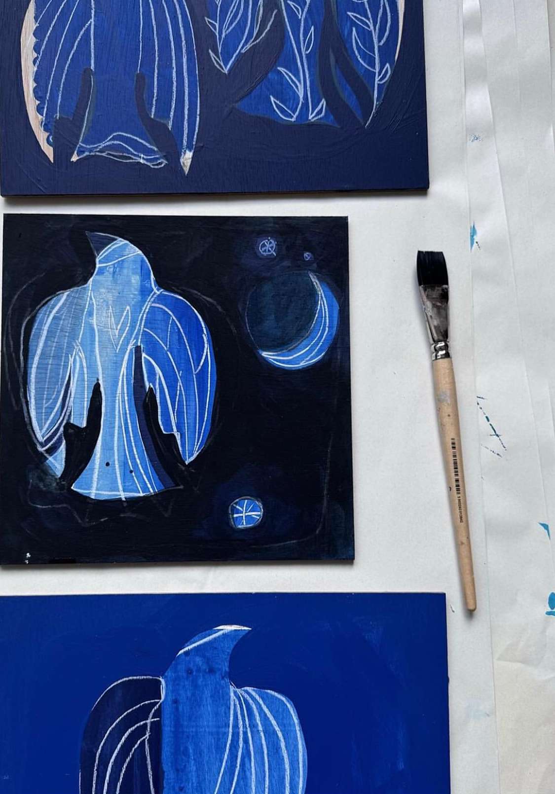 Three artworks of birds and plants painted in blue and white, displayed on a white surface next to a paintbrush.