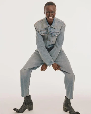 Smiling black model wearing a light blue denim jacket and matching jeans with black leather boots against a white backdrop.