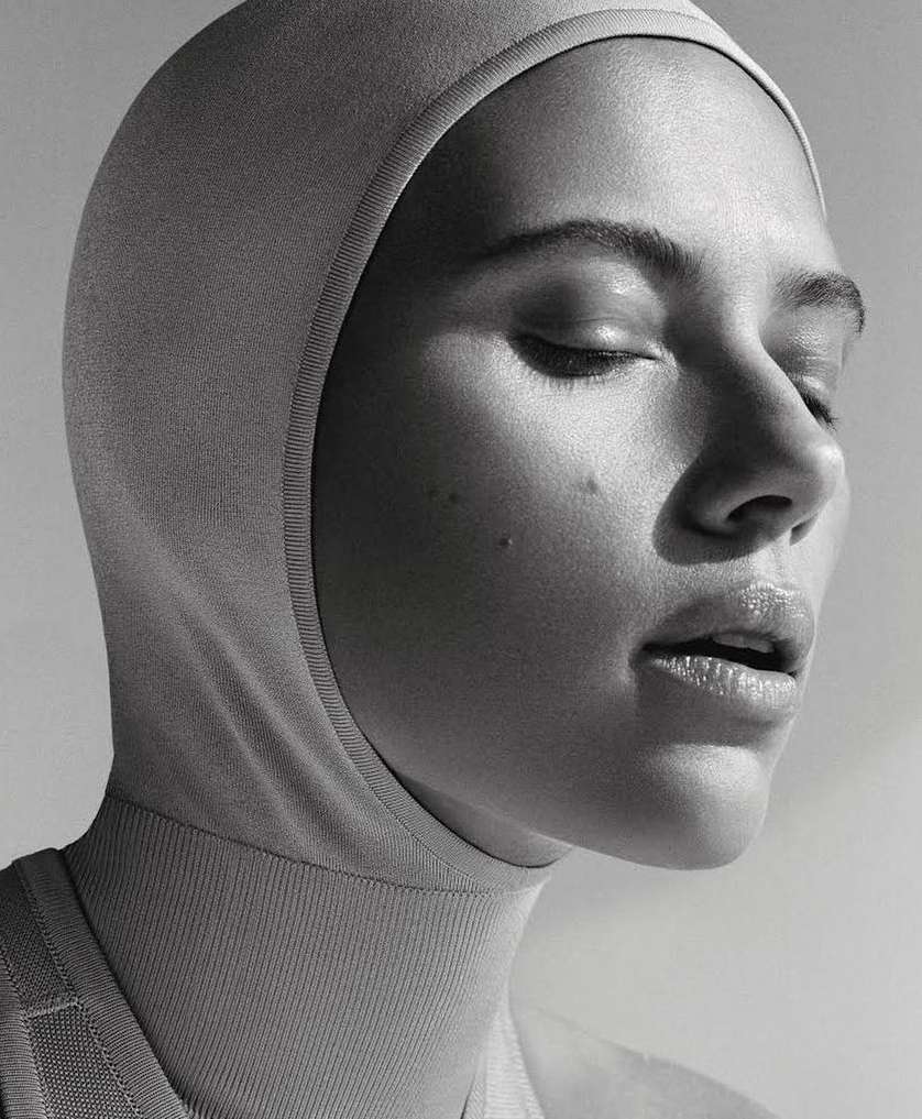 Monochrome portrait of a woman wearing a fitted hood covering her head and neck, with her eyes closed in soft lighting.