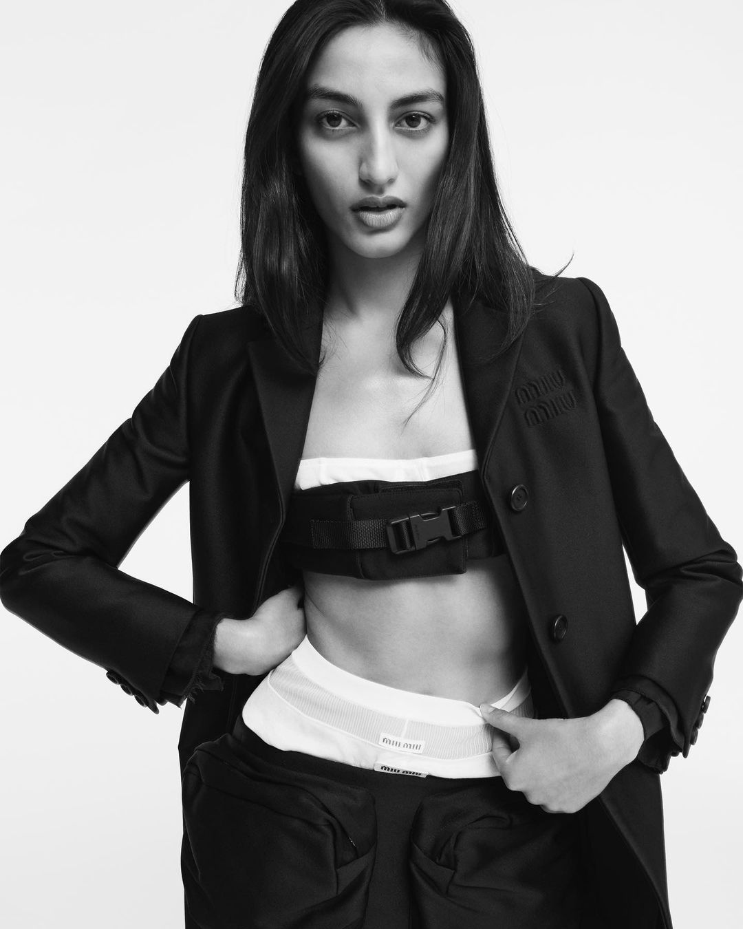 Studio shot of a model wearing a dark blazer and matching bottoms, with a white top and a dark utility belt on a light background.