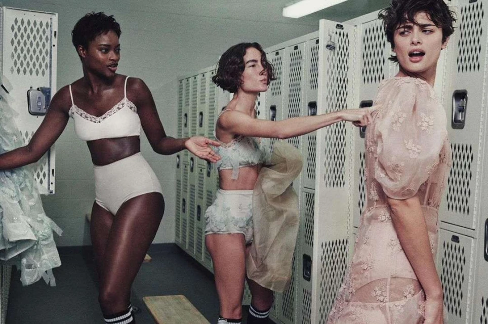 Three women in vintage-style lingerie and dresses pose in a locker room, creating a retro fashion editorial look.
