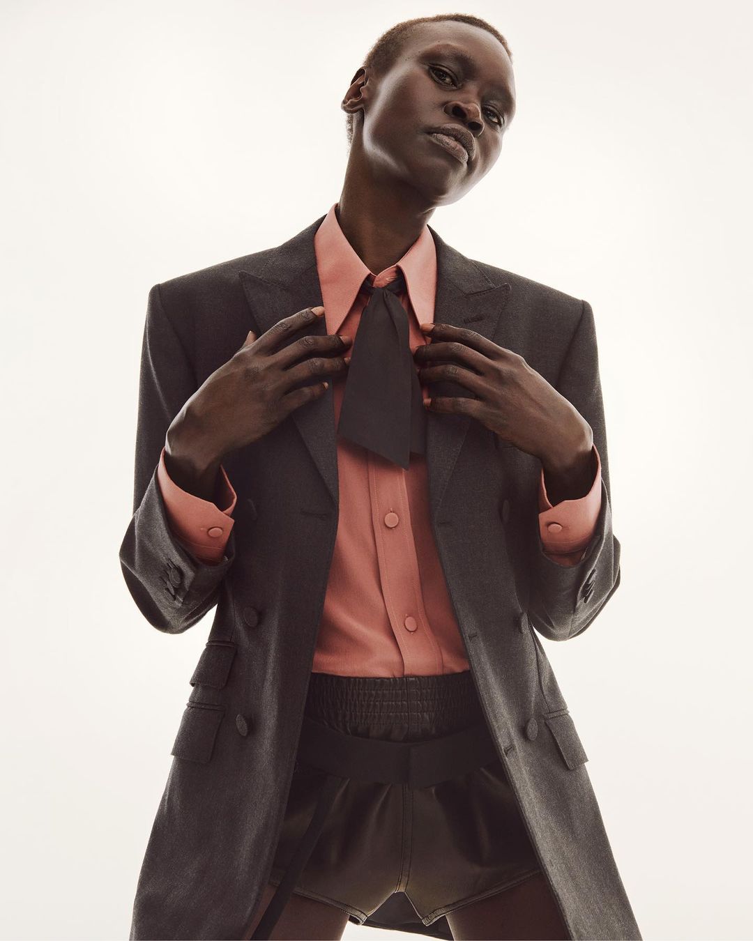 Model in dark grey blazer and peach shirt with black tie posing against a white background