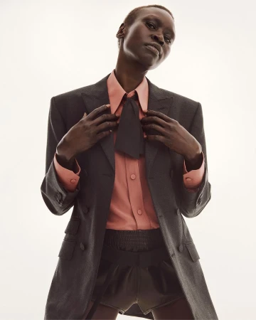 Model in dark grey blazer and peach shirt with black tie posing against a white background
