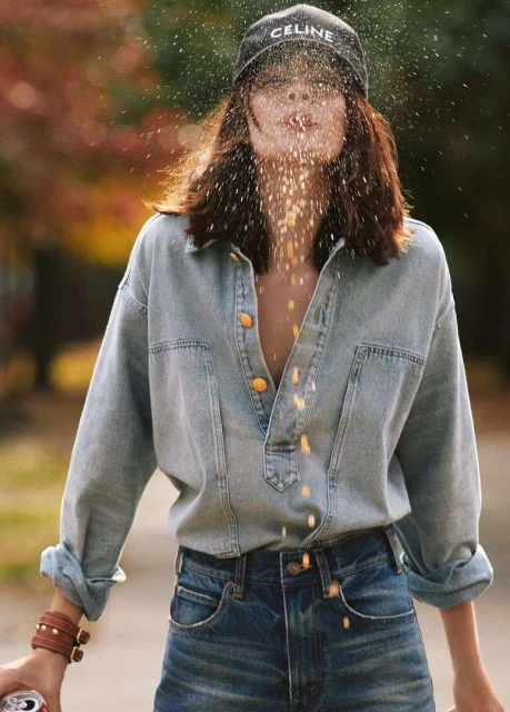 Woman in denim shirt and jeans throws grains into the air, wearing a Celine cap in an outdoor fall setting with vibrant foliage.