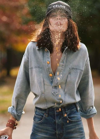 Woman in denim shirt and jeans throws grains into the air, wearing a Celine cap in an outdoor fall setting with vibrant foliage.