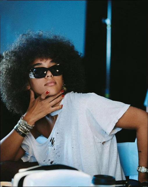 A woman with dark, curly hair and sunglasses poses indoors, wearing a ripped white t-shirt and jewelry, set against a blurred blue background.