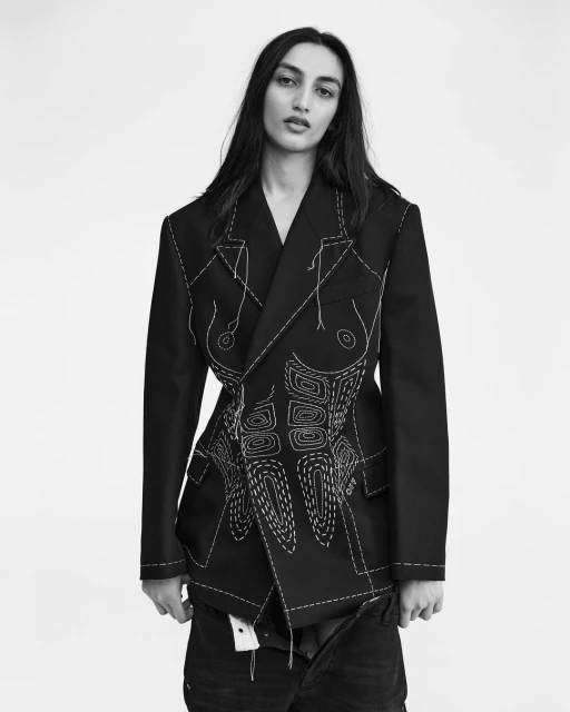 Monochrome portrait of an adult woman in a black jacket with white stitching details, standing indoors against a white background in a fashion shot.