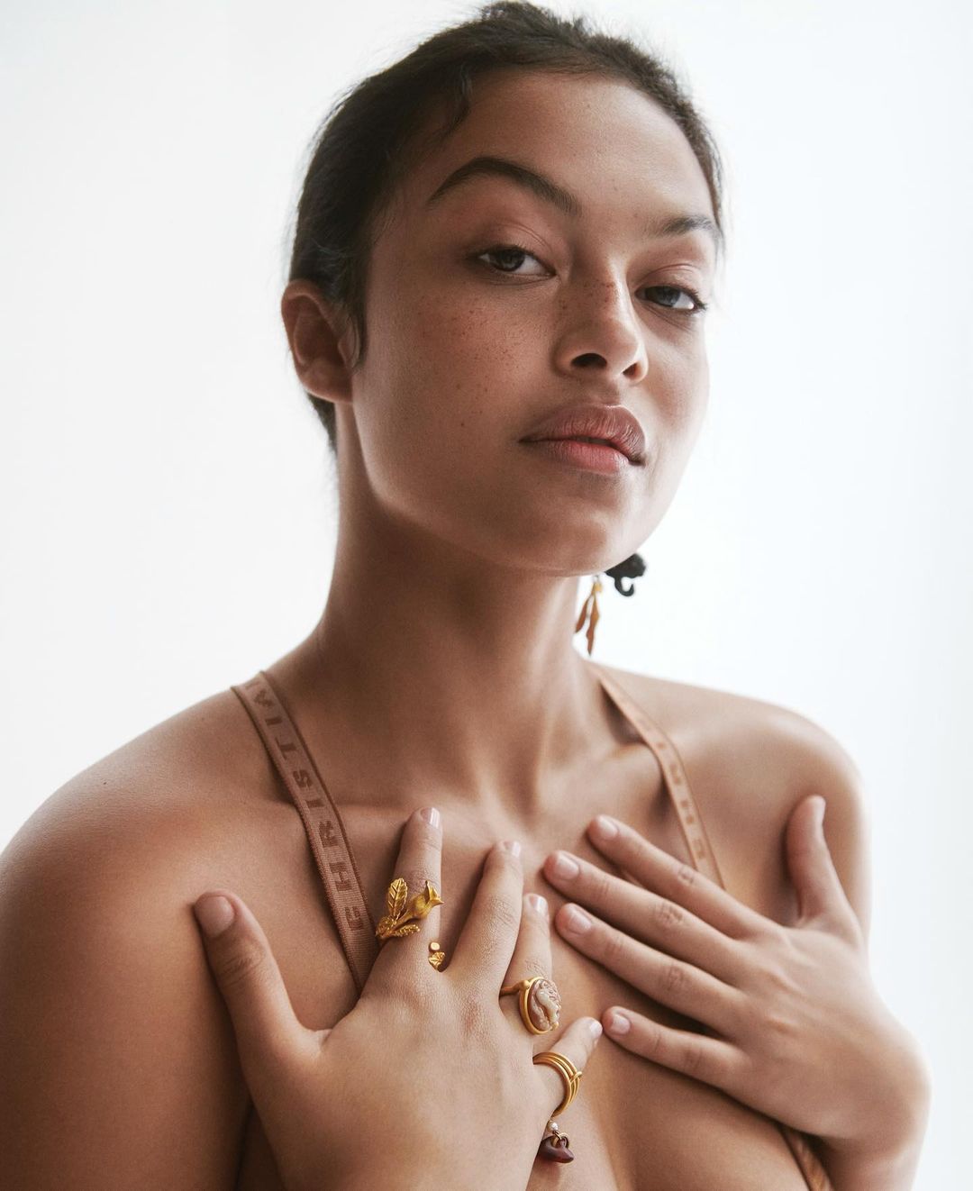 Portrait of an adult female with tanned skin and gold jewelry on her hands in a studio setting with a white background.