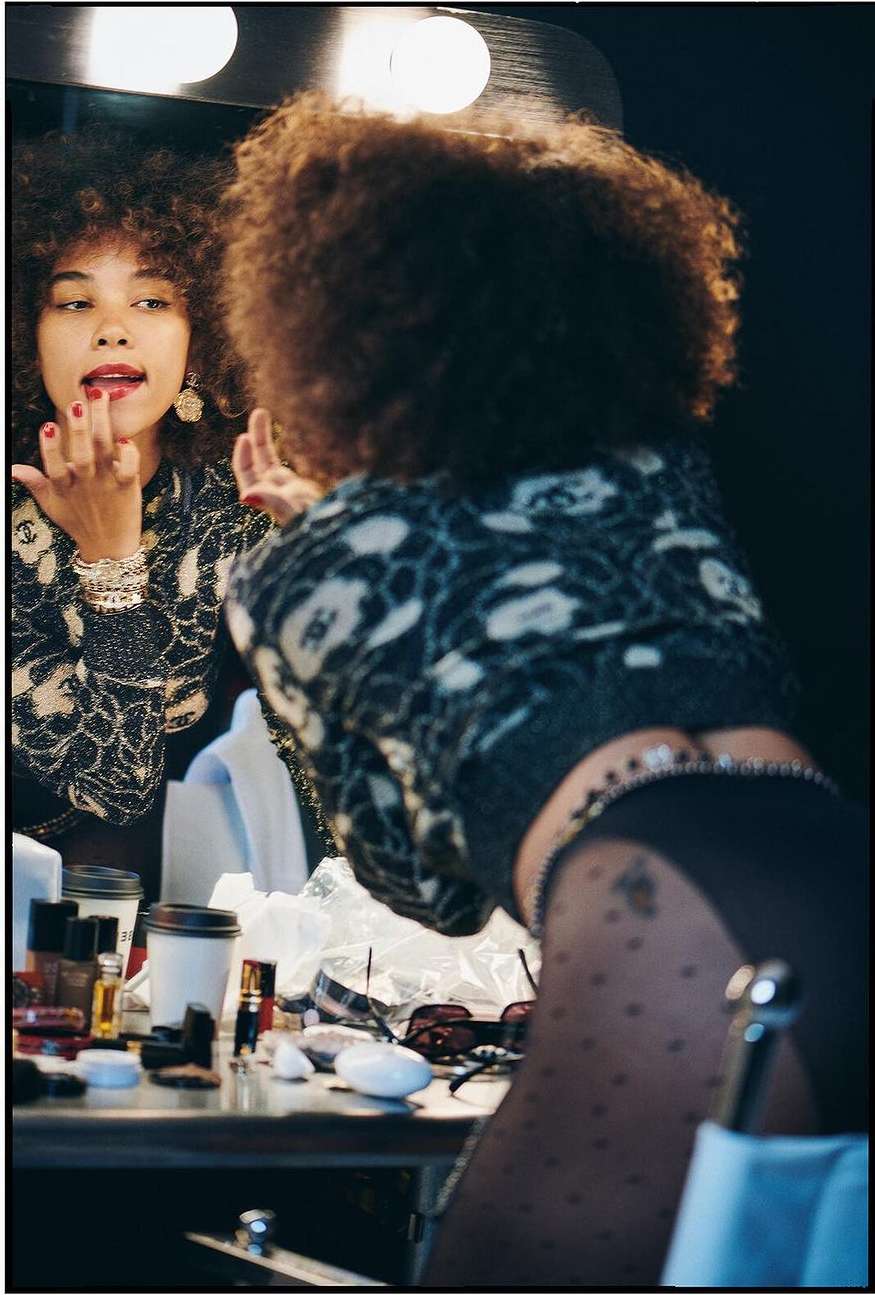 Woman with afro in a backstage dressing room applying red lipstick in the mirror. Makeup and other cosmetic items are on the table.