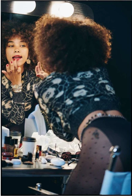 Woman with afro in a backstage dressing room applying red lipstick in the mirror. Makeup and other cosmetic items are on the table.