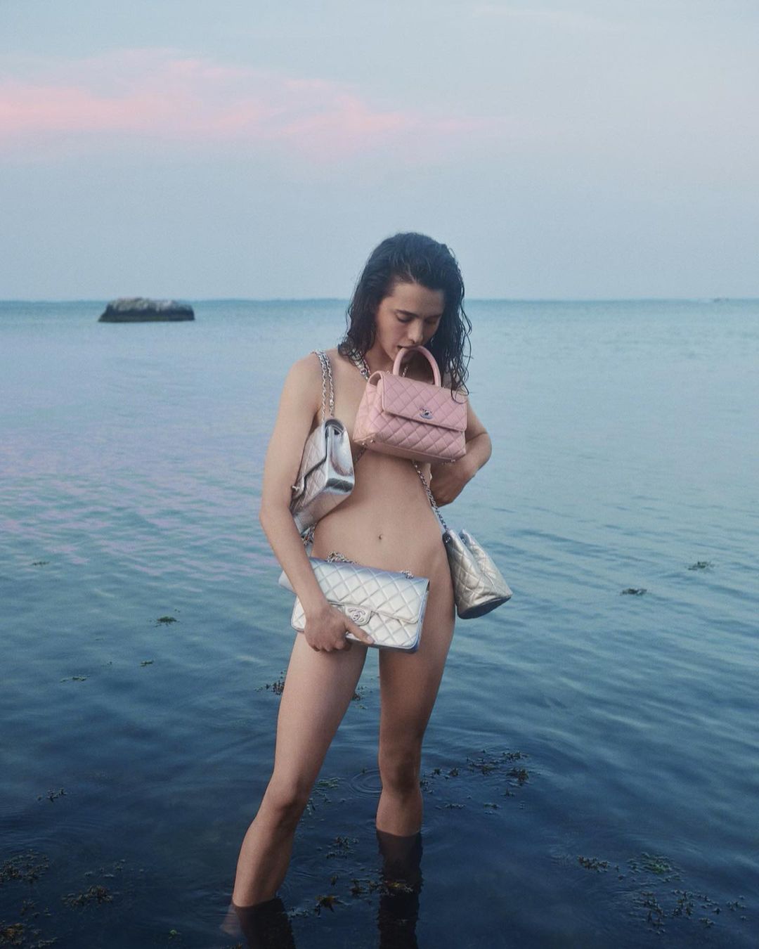 Woman standing in shallow ocean water holding multiple quilted designer handbags by photographer Cass Bird