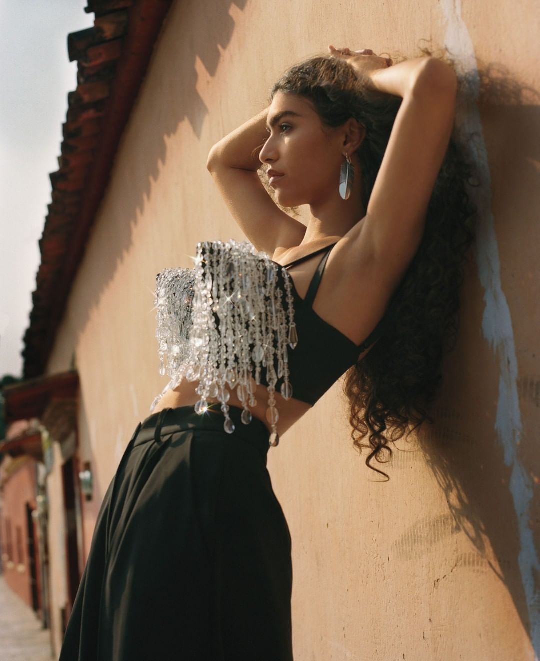 Fashionable woman with curly hair wearing a beaded top and leaning against a sunlit wall in an outdoor, urban setting.