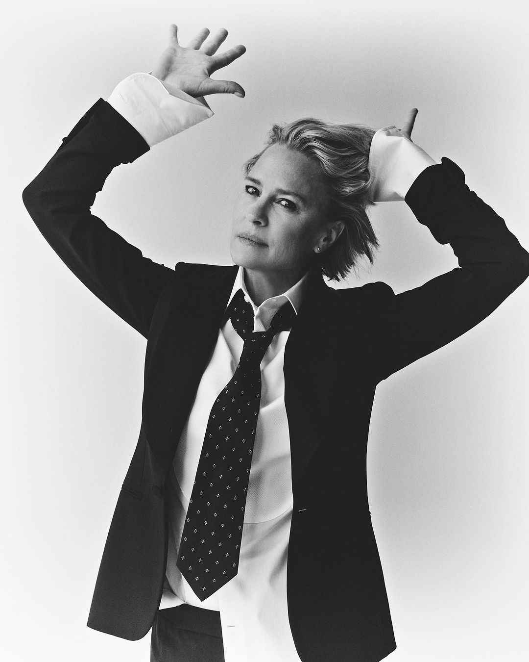 Black and white portrait of a blonde adult woman in a black suit, white shirt, and patterned tie posing against a white background.