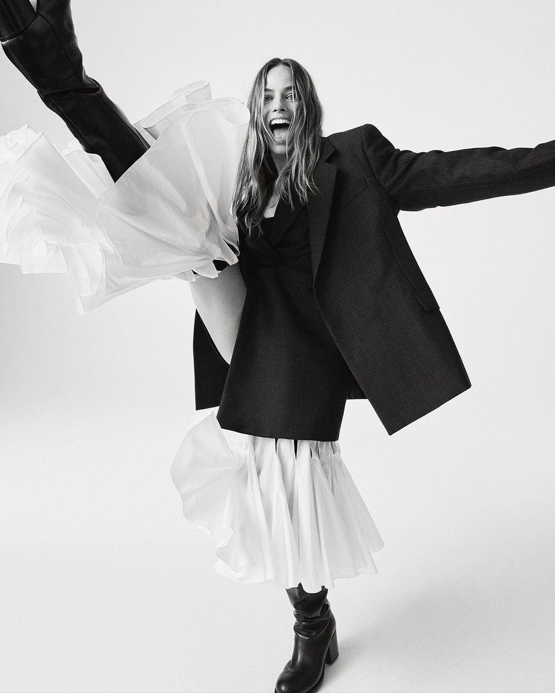 Monochrome full shot of an adult woman wearing a dark blazer, dress with a ruffled hem, and boots, striking a joyful pose in a studio.