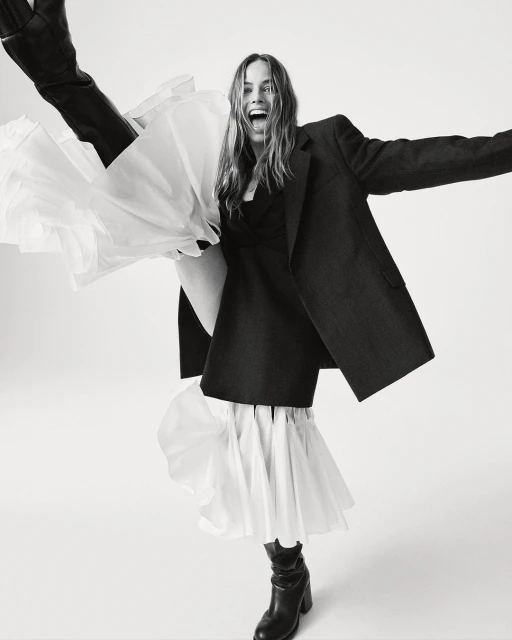 Monochrome full shot of an adult woman wearing a dark blazer, dress with a ruffled hem, and boots, striking a joyful pose in a studio.