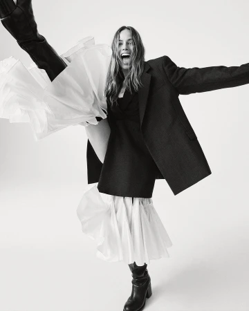 Monochrome full shot of an adult woman wearing a dark blazer, dress with a ruffled hem, and boots, striking a joyful pose in a studio.