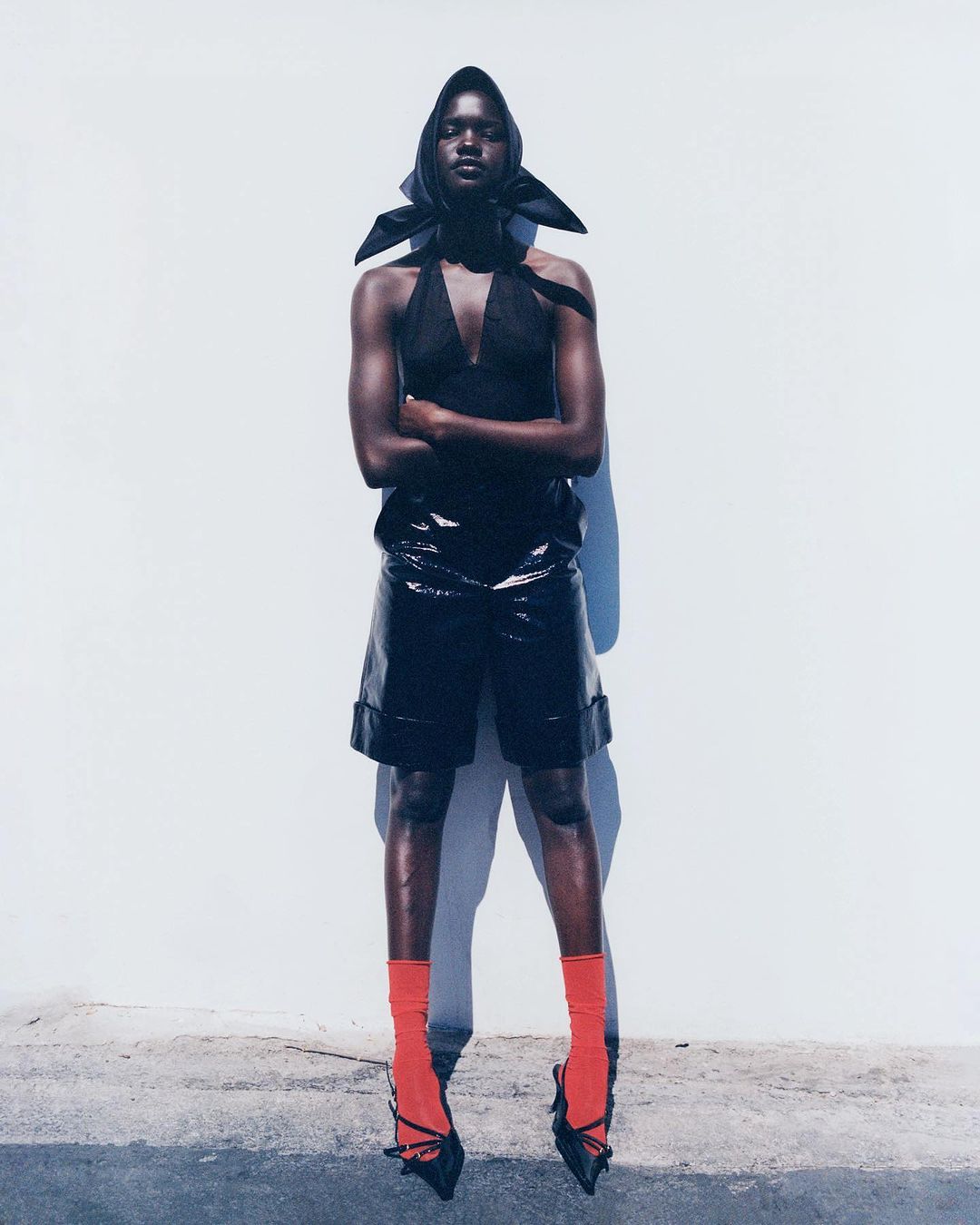 Fashion portrait of a Black woman in a black halter top, black patent leather shorts, orange socks, and black heels posing against a wall.