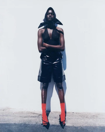 Fashion portrait of a Black woman in a black halter top, black patent leather shorts, orange socks, and black heels posing against a wall.