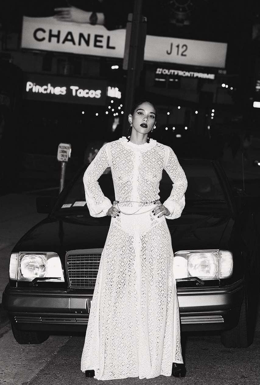 A woman in a white lace dress stands in front of a black car at night, framed by city lights and billboards. Fashion and urban setting.