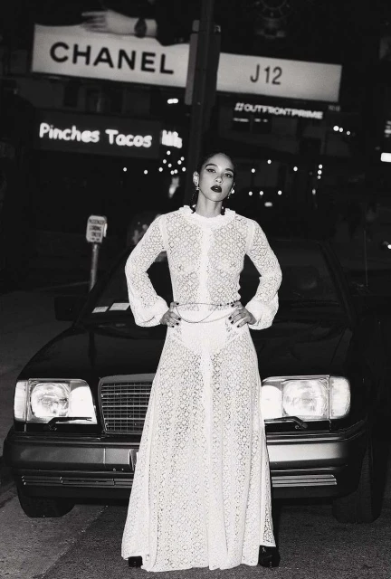 A woman in a white lace dress stands in front of a black car at night, framed by city lights and billboards. Fashion and urban setting.