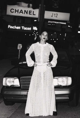 A woman in a white lace dress stands in front of a black car at night, framed by city lights and billboards. Fashion and urban setting.