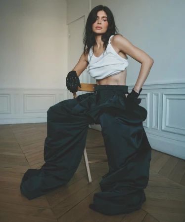 Brunette woman sitting on a chair in a white tank top and black gloves with wide leg pants in a light-filled interior.
