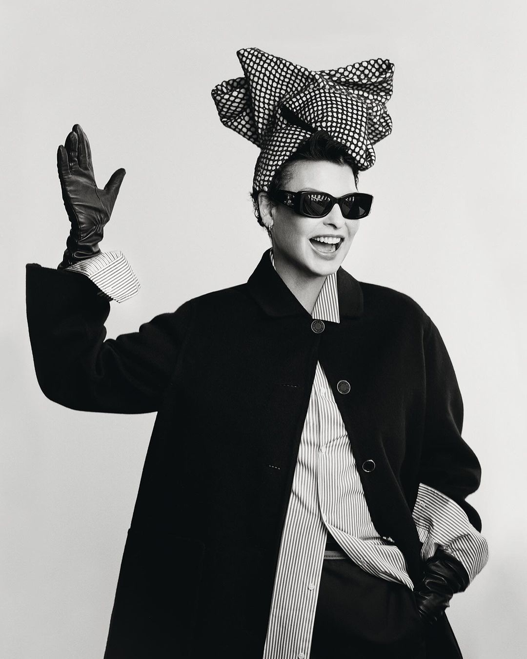 Model wearing dark sunglasses, striped shirt, black coat, leather gloves, and large patterned head wrap waving against white background.