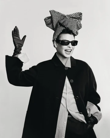 Model wearing dark sunglasses, striped shirt, black coat, leather gloves, and large patterned head wrap waving against white background.