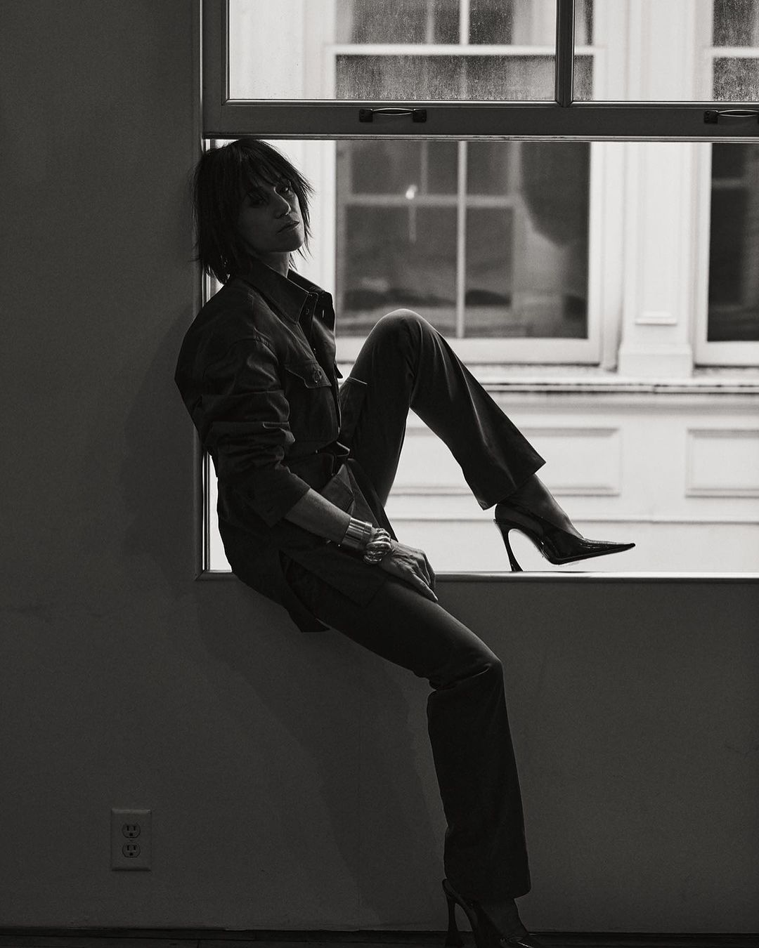 A woman in a jumpsuit and heels sits in a window in a black and white studio portrait; she wears a short, dark, shaggy hairstyle.