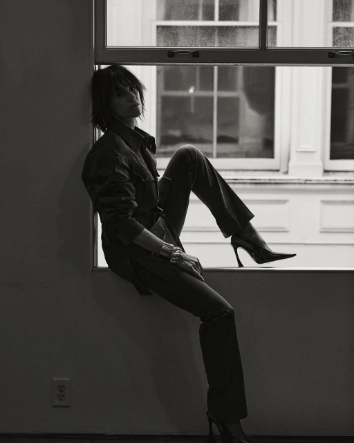 A woman in a jumpsuit and heels sits in a window in a black and white studio portrait; she wears a short, dark, shaggy hairstyle.