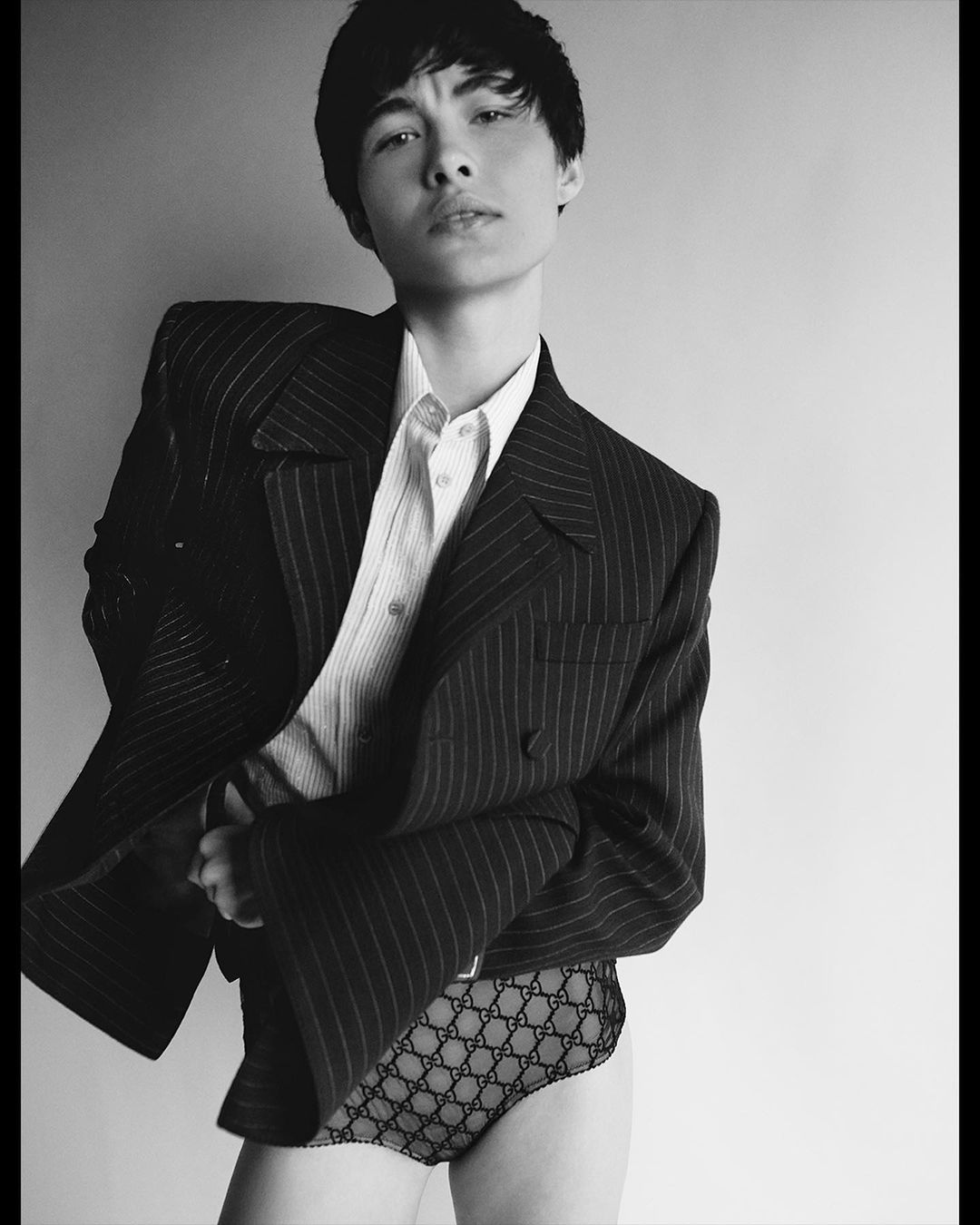 Black-and-white studio portrait of a young adult in a pinstripe blazer, white shirt, and designer underwear against a gradient backdrop.