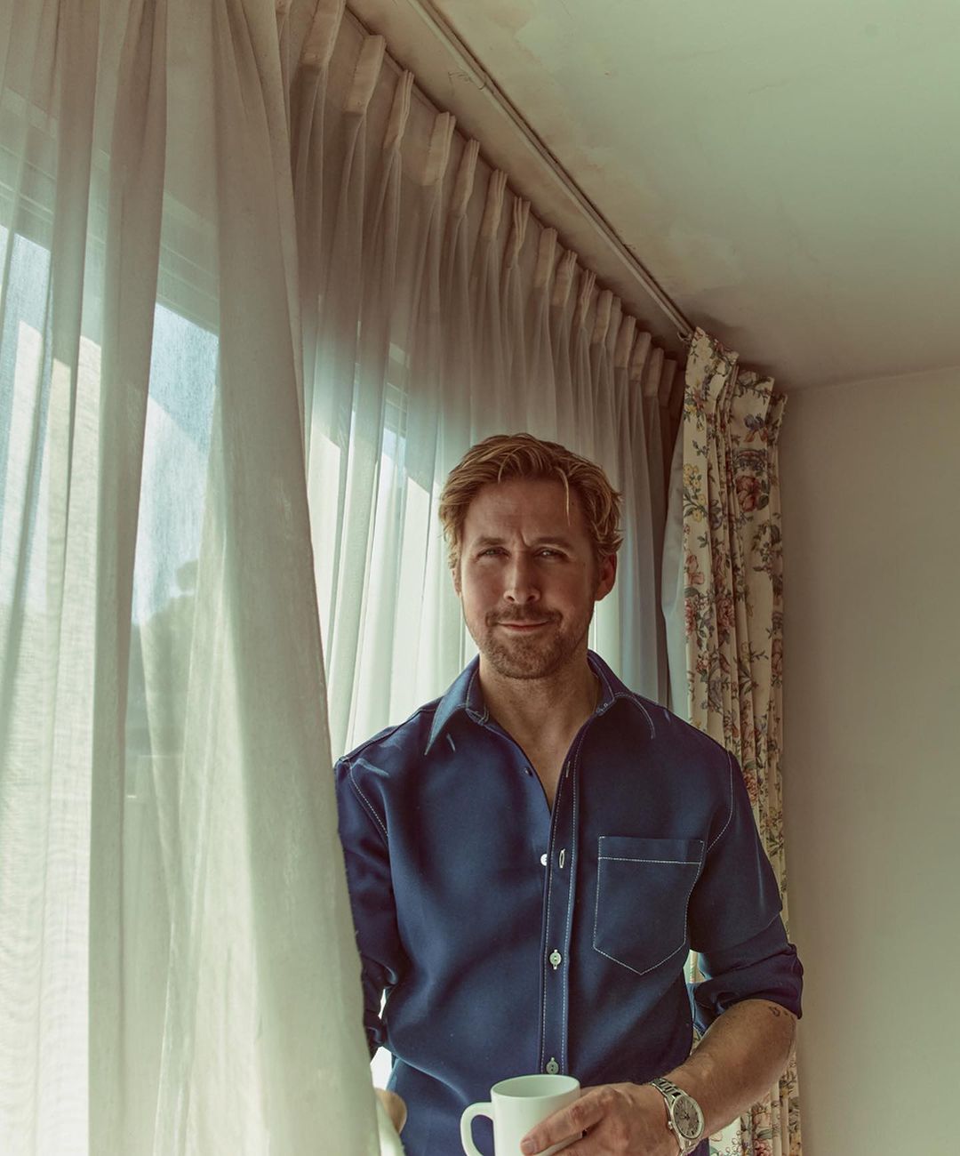 Blond adult male in a blue shirt poses near a window holding a white mug, with light coming through white curtains and a neutral wall.
