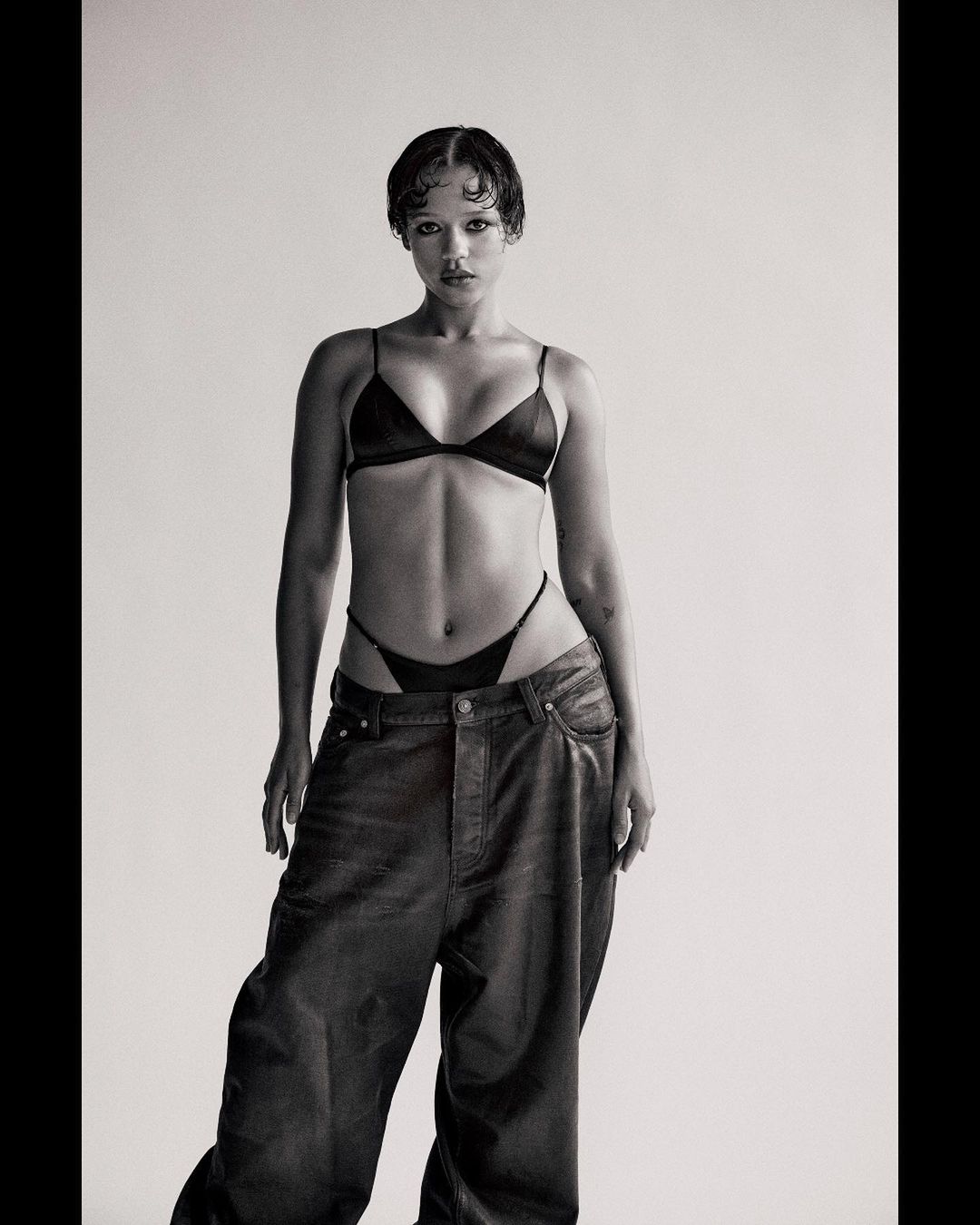 Monochrome studio shot of an adult female model in a black bra, thong, and baggy jeans, posing against a neutral backdrop with soft lighting.