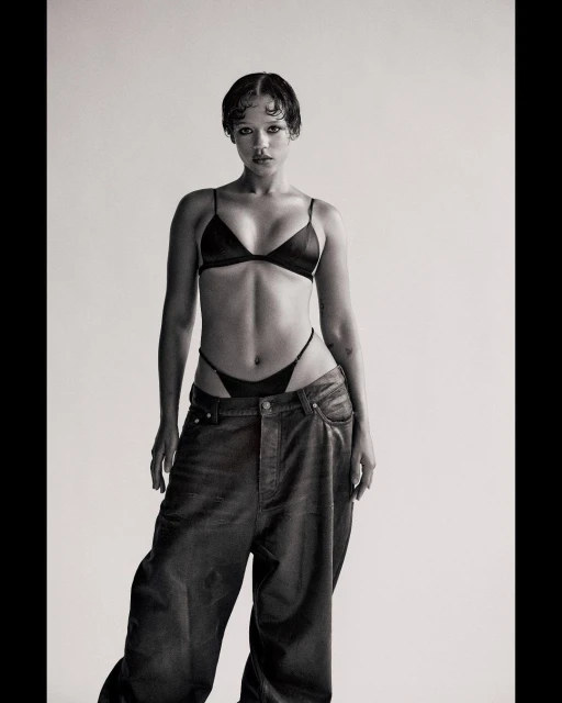 Monochrome studio shot of an adult female model in a black bra, thong, and baggy jeans, posing against a neutral backdrop with soft lighting.