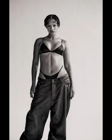 Monochrome studio shot of an adult female model in a black bra, thong, and baggy jeans, posing against a neutral backdrop with soft lighting.