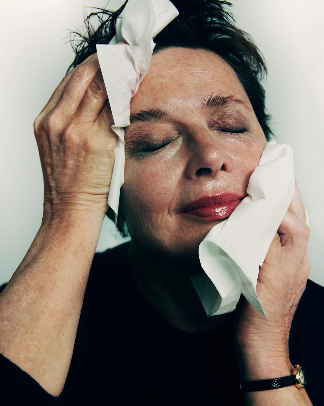 Close-up portrait of a woman with a black shirt, holding tissue to her face, with closed eyes, indoors, against a white backdrop.