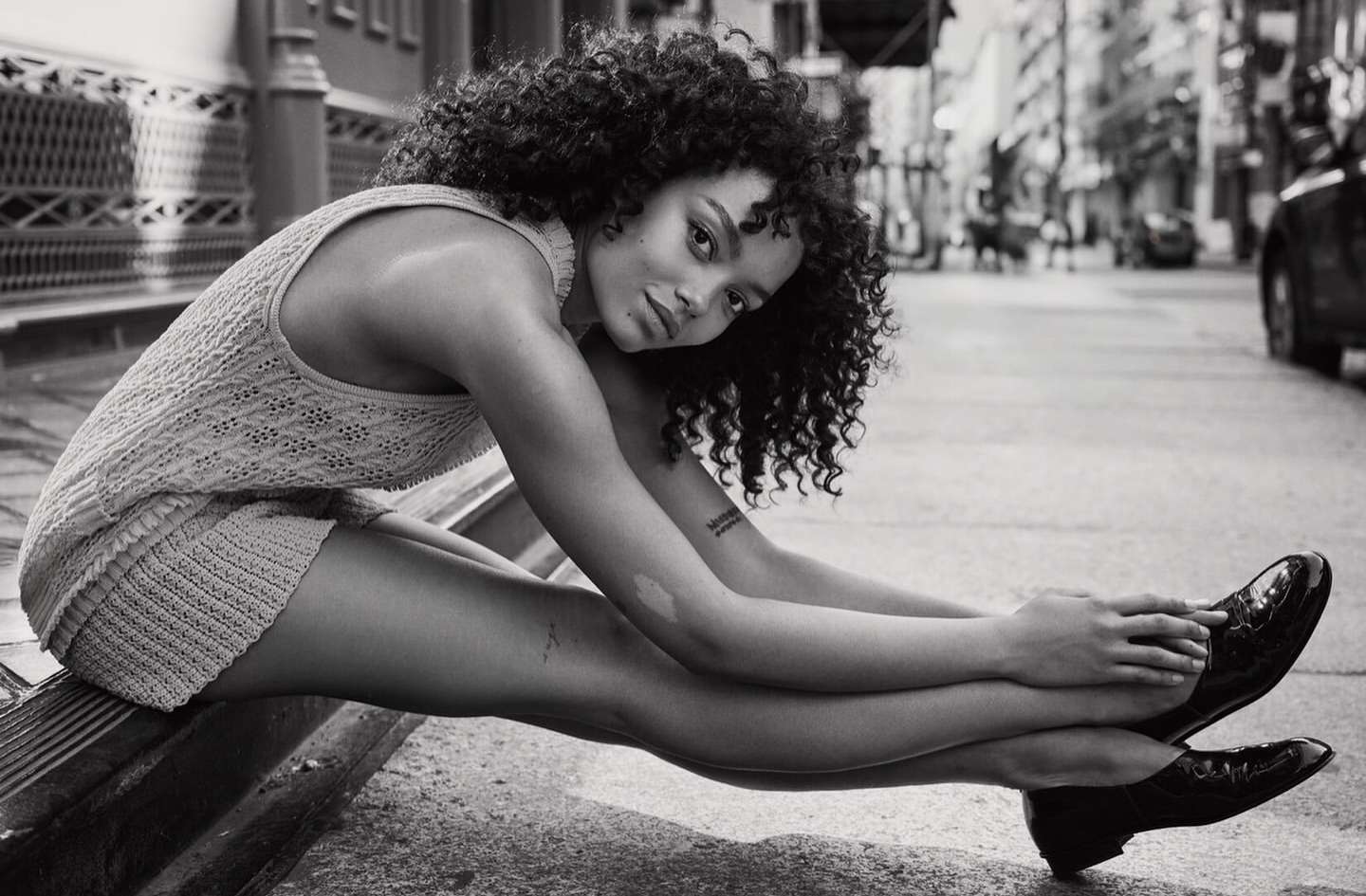 Black and white shot of a woman with curly hair wearing a knit outfit and black patent leather shoes sitting on a city street.