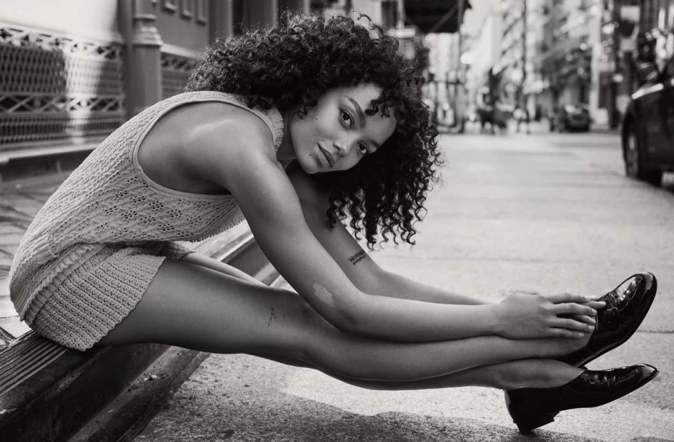 Black and white shot of a woman with curly hair wearing a knit outfit and black patent leather shoes sitting on a city street.