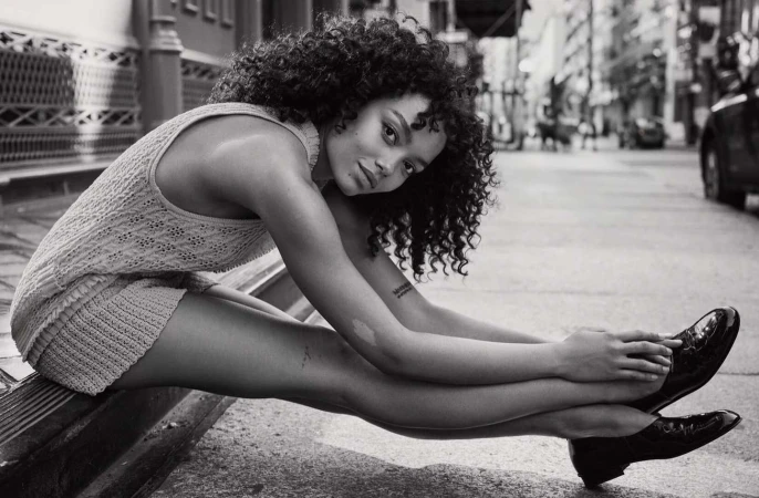 Black and white shot of a woman with curly hair wearing a knit outfit and black patent leather shoes sitting on a city street.