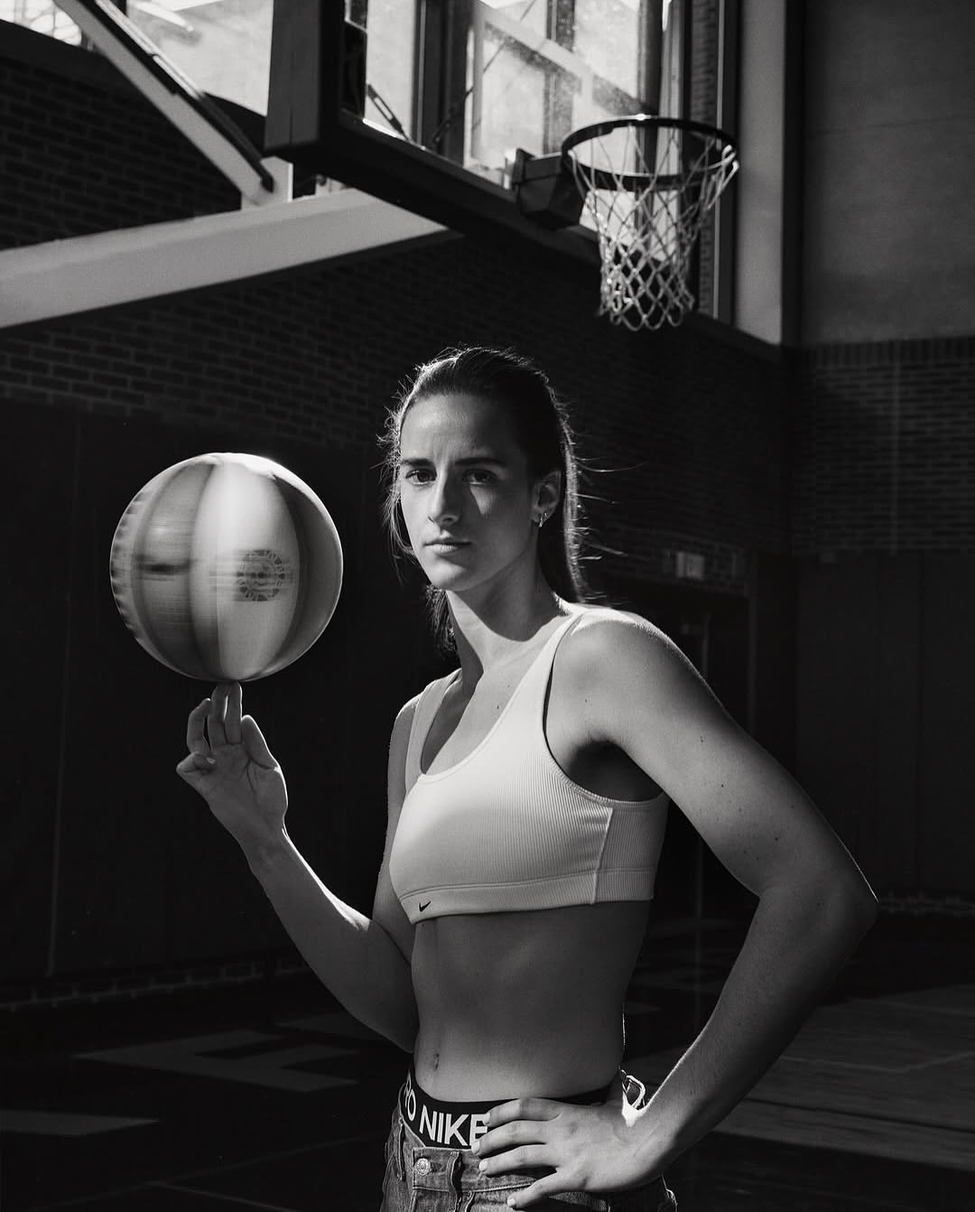 Athletic woman spinning a basketball on her finger in a gym. She's wearing athletic clothes with a Nike logo, and there's a hoop behind her.