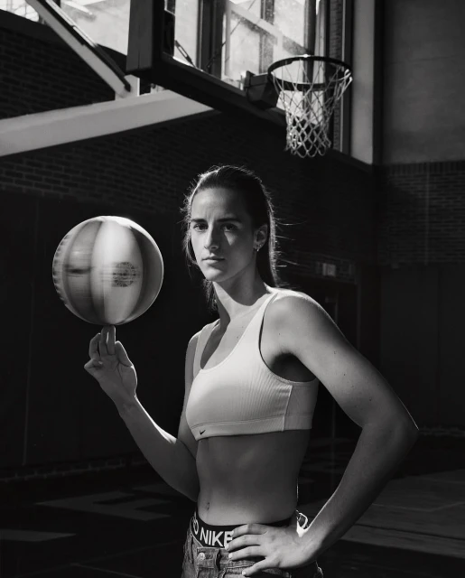 Athletic woman spinning a basketball on her finger in a gym. She's wearing athletic clothes with a Nike logo, and there's a hoop behind her.