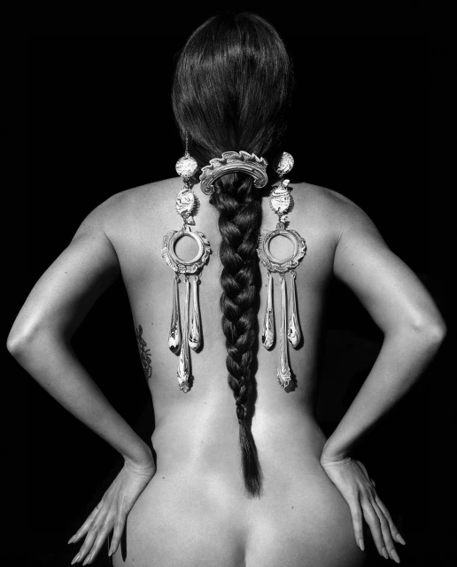 Back view of an adult female with long, dark hair styled in a braid, adorned with decorative metal ornaments and black background.