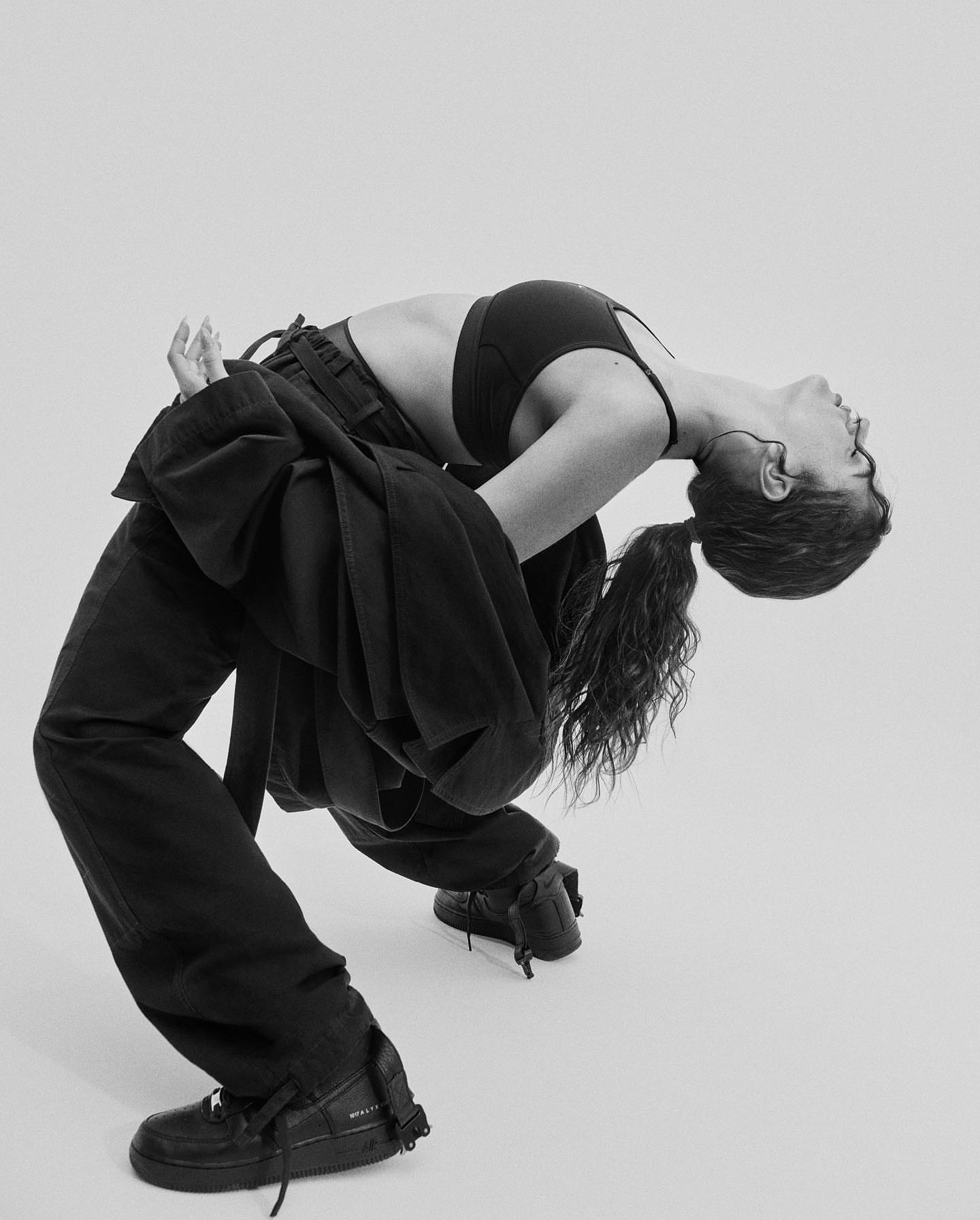 A woman bends backward in a black bra top, pants, and sneakers against a white backdrop in a studio setting, showcasing fashion and fitness.