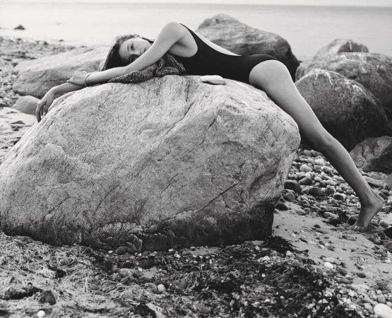 Model in black one-piece swimsuit draped over a large boulder on a rocky beach by photographer Cass Bird