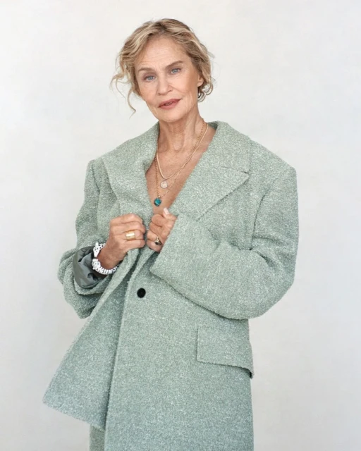 Older woman poses in a light green textured blazer with jewelry against a white backdrop. Fashion editorial shot, medium framing.