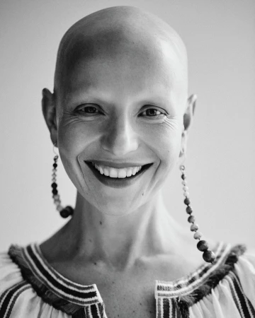 Close-up portrait of a smiling, bald adult woman with long, beaded earrings, wearing a striped blouse, against a neutral background.