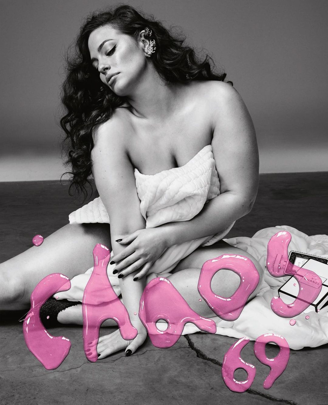 Ashley Graham poses wrapped in a towel next to pink, fluid typography and a Chanel box on a grey surface in an indoor studio setting.
